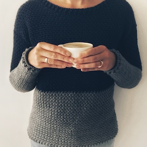 May include: A person wearing a dark gray and light gray striped sweater with a scoop neck. The person is holding a white mug with a light brown beverage.