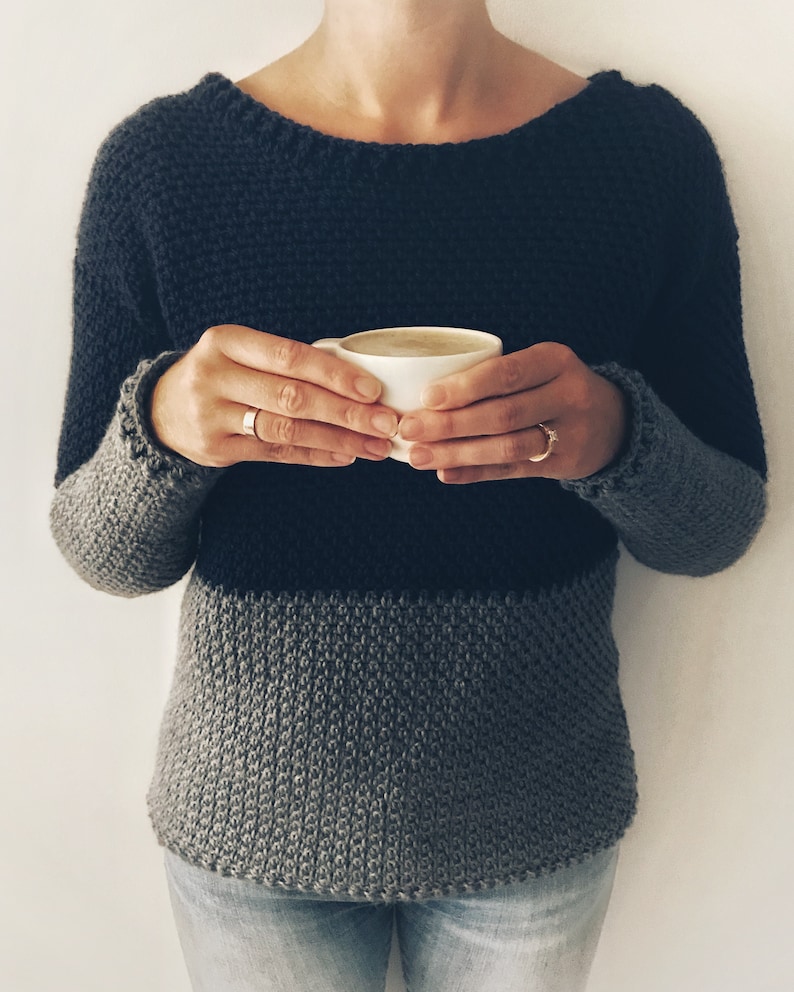 May include: A person wearing a dark gray and light gray striped sweater with a scoop neck. The person is holding a white mug with a light brown beverage.