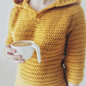 May include: A woman wearing a yellow crocheted hooded sweater holds a white mug of coffee.