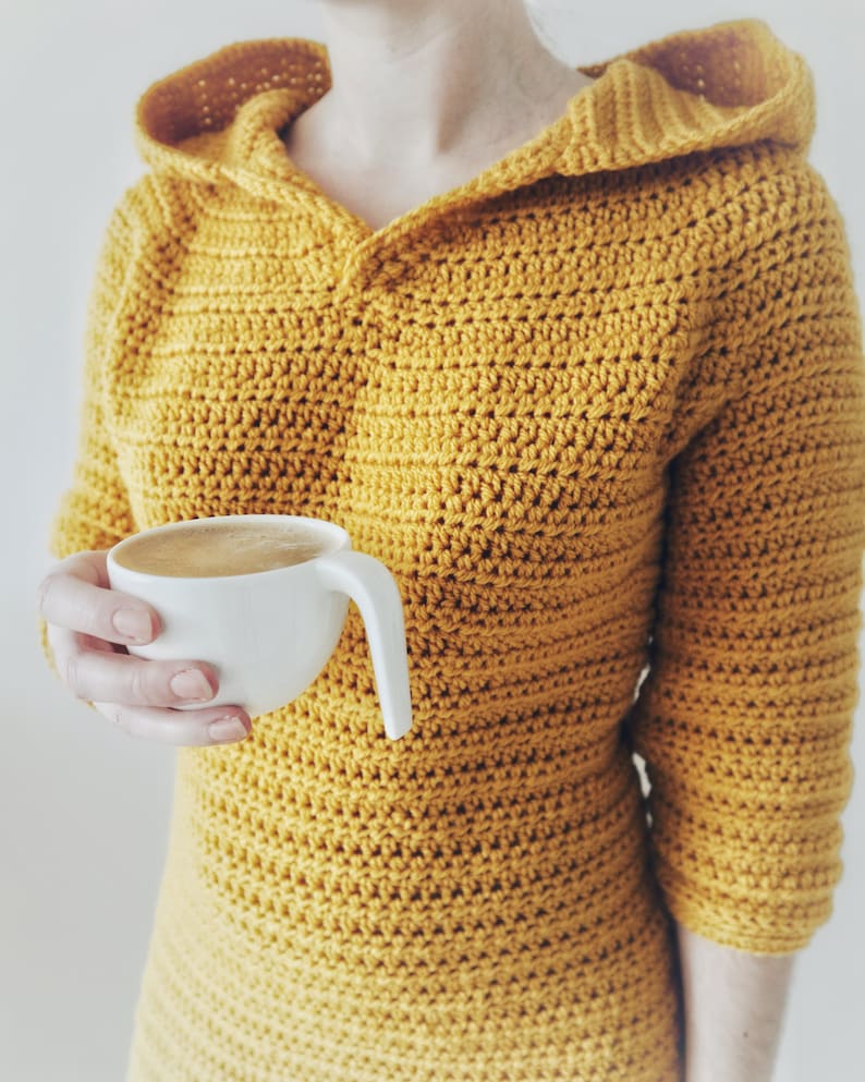 May include: A woman wearing a yellow crocheted hooded sweater holds a white mug of coffee.