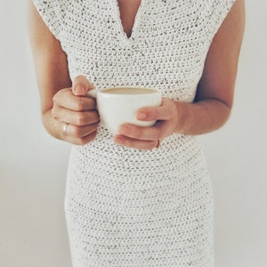 May include: A white crocheted dress with a v-neck and short sleeves. The person is holding a white cup of coffee.