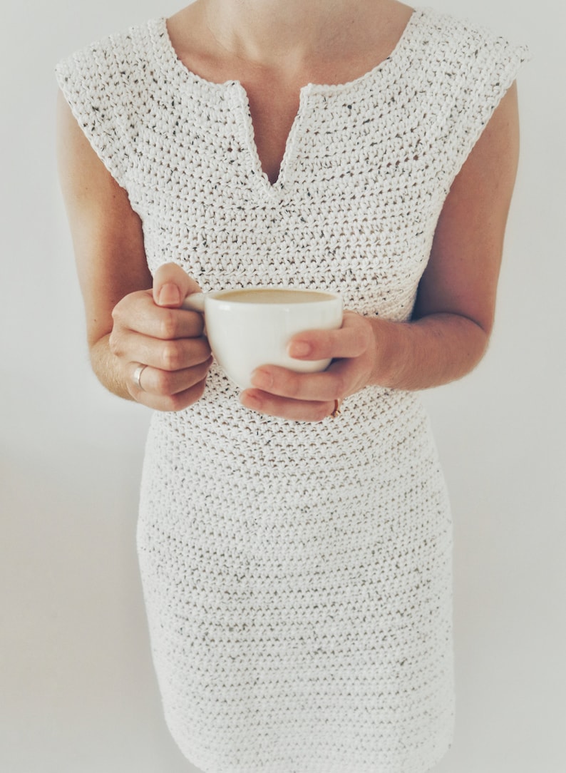 May include: A white crocheted dress with a v-neck and short sleeves. The person is holding a white cup of coffee.