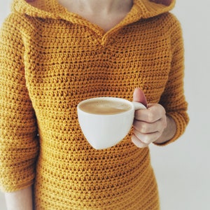May include: A person wearing a mustard yellow crocheted hooded sweater holds a white mug of coffee.
