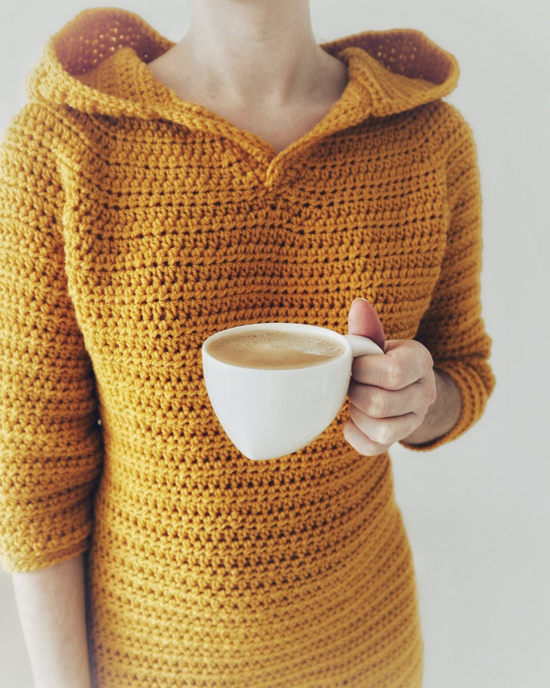 May include: A person wearing a mustard yellow crocheted hooded sweater holds a white mug of coffee.