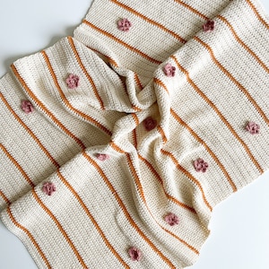 May include: A white and orange striped crocheted baby blanket with pink flower embellishments.
