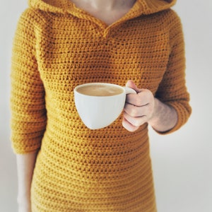 May include: A person wearing a mustard yellow crocheted hooded dress, holding a white mug filled with coffee.