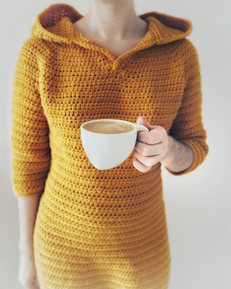 May include: A person wearing a mustard yellow crocheted hooded dress, holding a white mug filled with coffee.