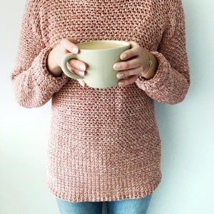 May include: A woman wearing a pink knitted sweater with a light beige mug in her hands.
