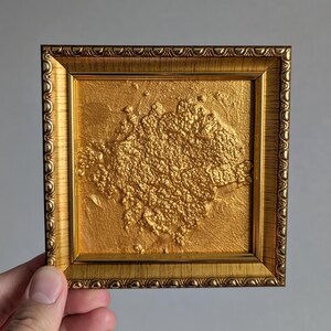 May include: A small, square, gold-colored framed artwork. The artwork inside the frame has a textured, raised surface with a gold finish. The frame itself has a detailed, ornate design.