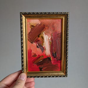 May include: Small framed abstract painting with a gold ornate frame. The artwork features bold brushstrokes in shades of red, brown, and white on a red background. The painting is held by a hand against a gray backdrop.