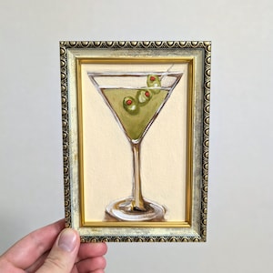 Może przedstawiać: Oprawiony obraz koktajlu Martini z trzema oliwkami na wykałaczce. Grafika przedstawia złotą ramę z dekoracyjną obwódką. Kieliszek do martini wypełniony jest zielonym płynem i stoi na jasnobeżowym tle.