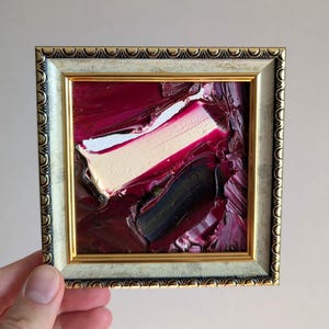 May include: A small square painting featuring thick, textured oil paint strokes in deep burgundy, dark purple, and a prominent diagonal streak of cream white. The artwork is encased in a decorative, ornate silver and gold-toned frame.