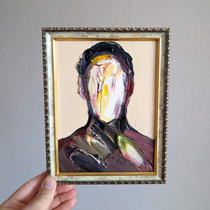 May include: Oil painting of a faceless figure in a decorative gold frame. The artwork features thick brushstrokes in shades of black, white, yellow, and burgundy. The background is a light beige color, and the frame has an ornate design.