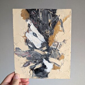 Könnte beinhalten: Ein rechteckiges Leinwandgemälde mit dicken, strukturierten Impasto-Farbschichten in einer Palette aus Creme, Schwarz, Weiß und Metallic-Gold. Die Komposition zeigt abstrakte Pinselstriche und Spachtelaufträge.
