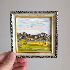 Pequeña pintura al óleo de Montana enmarcada, cielo azul y blanco texturizado, montañas escarpadas de color marrón grisáceo, campos vibrantes de color amarillo verdoso que evocan colinas onduladas