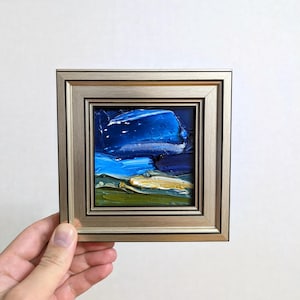 May include: Small square oil painting in a gold-colored frame. The artwork depicts a landscape with a dark blue sky, lighter blue water, and green and yellow land. The painting is held by a hand.