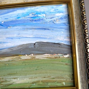 May include: Oil painting of a landscape with a mountain, green field, and blue sky with clouds. The painting is in a gold and off-white ornate frame. The artwork has visible brushstrokes.