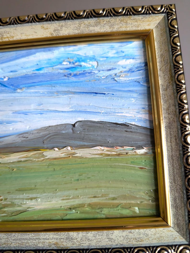 May include: Oil painting of a landscape with a mountain, green field, and blue sky with clouds. The painting is in a gold and off-white ornate frame. The artwork has visible brushstrokes.