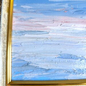 Op de afbeelding: Een olieverfschilderij van een zeegezicht met een blauwe lucht en water, en een roze en witte horizon. Het schilderij zit in een gouden lijst met een cr&egrave;mekleurige rand.