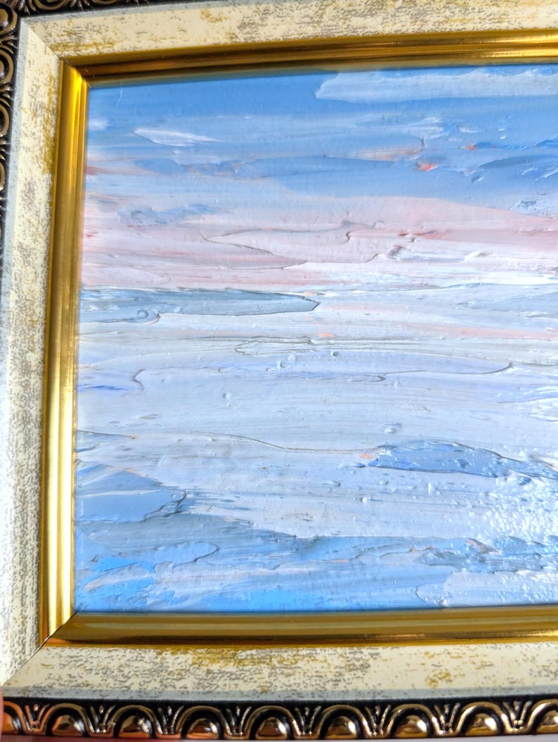 Op de afbeelding: Een olieverfschilderij van een zeegezicht met een blauwe lucht en water, en een roze en witte horizon. Het schilderij zit in een gouden lijst met een cr&egrave;mekleurige rand.