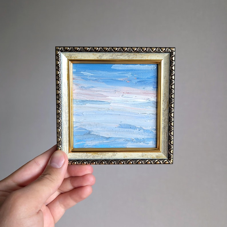 May include: Small framed oil painting depicting a sky scene with shades of blue and pink. The artwork is encased in a decorative gold and cream-colored frame, adding a touch of elegance to the miniature landscape.
