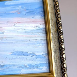 Op de afbeelding: Een olieverfschilderij van een zeegezicht in een goud-zwarte, sierlijke lijst. Het schilderij toont blauwe, witte en roze tinten, die een lucht- en waterlandschap uitbeelden. De lijst heeft een verweerde, vintage uitstraling.