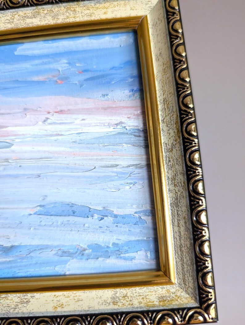 Op de afbeelding: Een olieverfschilderij van een zeegezicht in een goud-zwarte, sierlijke lijst. Het schilderij toont blauwe, witte en roze tinten, die een lucht- en waterlandschap uitbeelden. De lijst heeft een verweerde, vintage uitstraling.