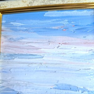 Op de afbeelding: Een olieverfschilderij van een zeegezicht met een textuur, impasto-stijl. Het kunstwerk heeft een palet van blauw, wit en vleugjes roze, omlijst door een gouden en cr&egrave;mekleurige sierlijke lijst. De penseelstreken cre&euml;ren een gevoel van beweging en diepte.