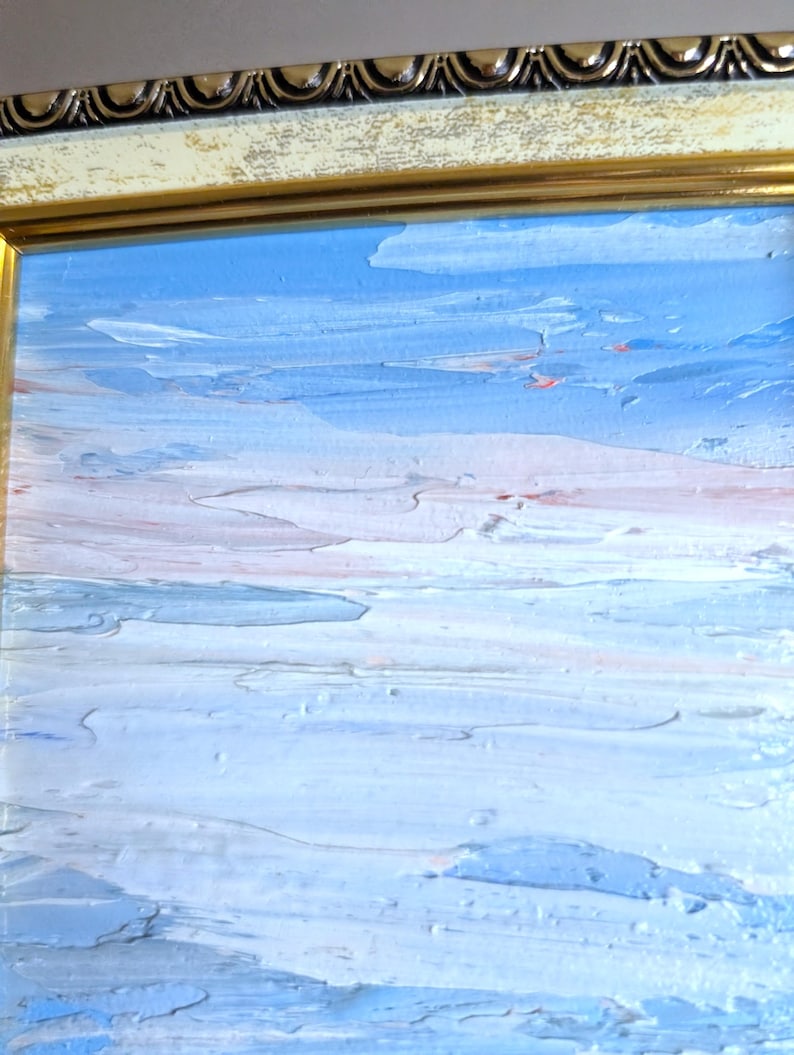 Op de afbeelding: Een olieverfschilderij van een zeegezicht met een textuur, impasto-stijl. Het kunstwerk heeft een palet van blauw, wit en vleugjes roze, omlijst door een gouden en cr&egrave;mekleurige sierlijke lijst. De penseelstreken cre&euml;ren een gevoel van beweging en diepte.
