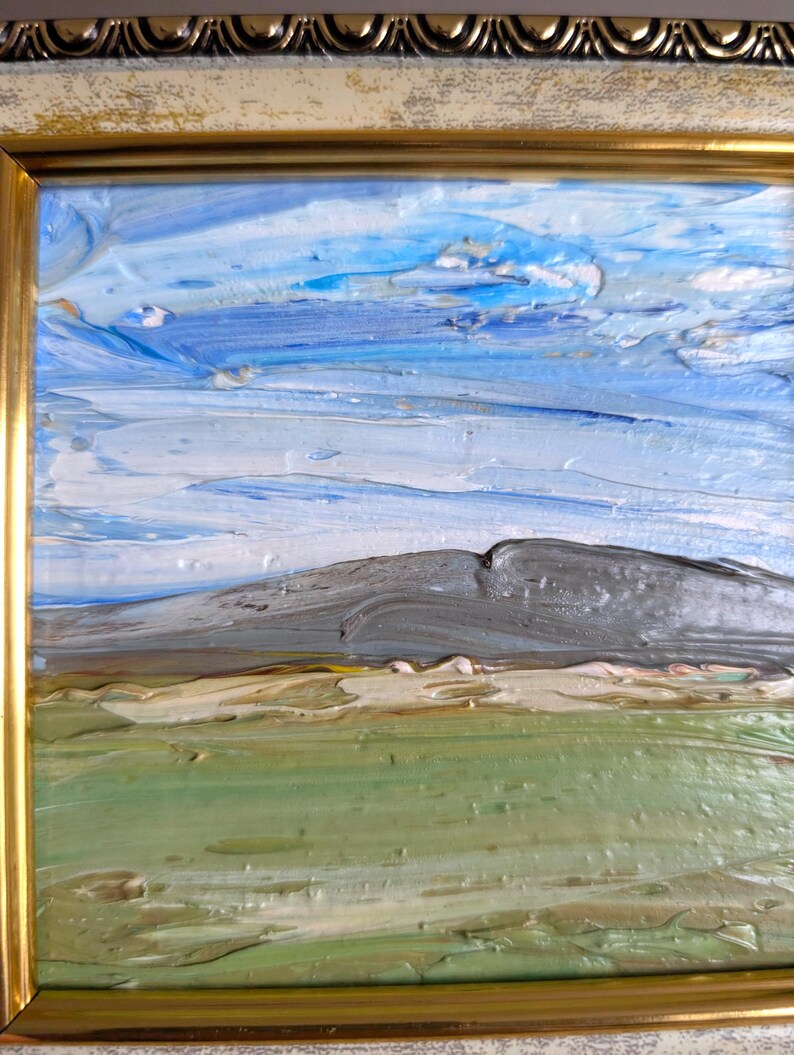 May include: Impasto oil painting of a landscape with a mountain, sky, and field. The sky is a mix of blues and white, the mountain is gray, and the field is green and tan. The painting is in a gold frame.