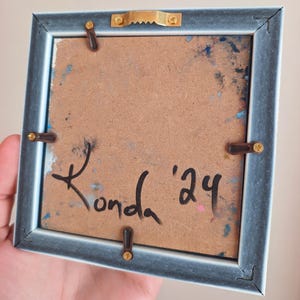 May include: Back of a small, square framed artwork. The frame is a muted blue-gray color. The artwork's backing is a light brown with paint splatters in blue and pink. The artist's signature, "Konda '24", is written in black ink.
