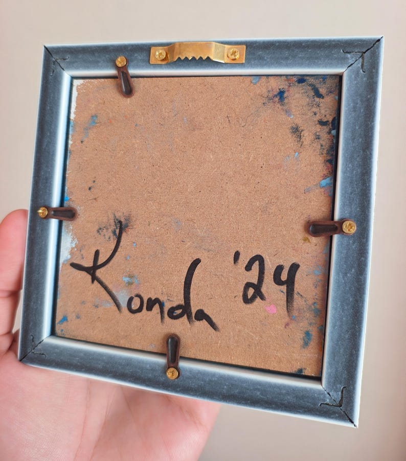 May include: Back of a small, square framed artwork. The frame is a muted blue-gray color. The artwork's backing is a light brown with paint splatters in blue and pink. The artist's signature, "Konda '24", is written in black ink.