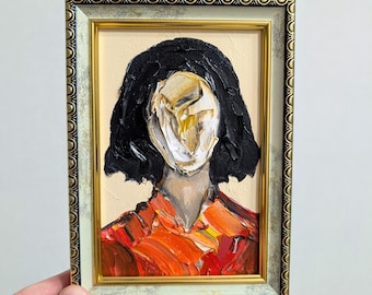 May include: An oil painting in a decorative gold and silver frame featuring a portrait with thick, textured brushstrokes. The subject has black hair, a white and gold abstract face, and wears a shirt with vibrant orange and red paint strokes.