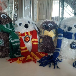 Könnte beinhalten: Vier Plüsch-Eulenspielzeuge, jedes mit einem andersfarbigen gestrickten Schal mit einem Hogwarts-Hauswappen. Die Eulen sind braun, weiß und grau.