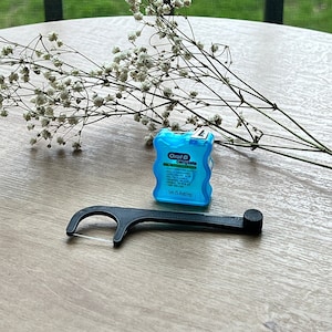 May include: A blue Oral-B Complete Floss Picks dispenser with a black floss pick on a wooden table. The dispenser has the text "Oral-B Complete Floss Picks" and "5m (5.4yd/45m)".