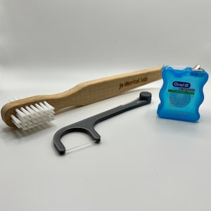 May include: A bamboo toothbrush with white bristles and the text js dental lab, a black plastic dental floss pick, and a blue Oral-B Complete SatinFloss container. These three oral hygiene items are arranged together on a plain surface.