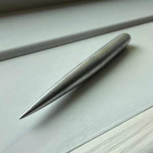 Op de afbeelding: Een zilverkleurige metalen priem met een puntige punt, liggend op een wit oppervlak. De priem heeft een glad, cilindrisch lichaam dat taps toeloopt naar een scherpe punt. De achtergrond is een wit geschilderd oppervlak.