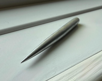 May include: A silver metal awl with a pointed tip, resting on a white surface. The awl has a smooth, cylindrical body that narrows to a sharp point. The background is a white painted surface.