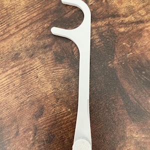 May include: A white dental floss pick with a curved head and a handle. The pick has a small, round, textured pad at the base of the handle. The object is on a brown wooden surface.