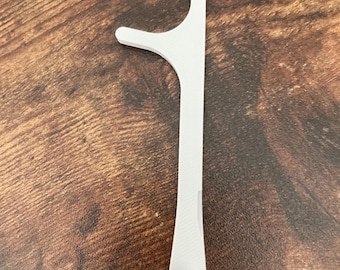 May include: A white dental floss pick with a curved head and a handle. The pick has a small, round, textured pad at the base of the handle. The object is on a brown wooden surface.