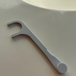 May include: A light gray, 3D-printed dental floss pick featuring a curved, hook-shaped head for holding floss and a textured, circular grip at the opposite end of the handle, designed for oral hygiene and cleaning between teeth.