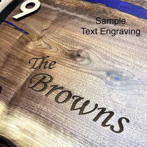 Peut inclure: Gros plan sur un cadran d'horloge en bois avec le chiffre 9 de couleur claire. L'horloge présente un motif de rivière en résine bleue et le texte gravé "The Browns" et "Sample Text Engraving". Le grain du bois est visible.
