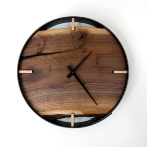 May include: A circular wooden wall clock with a dark metal frame. The clock face is made of natural wood with a visible grain. Black clock hands and small wooden accents. The clock is approximately 30 cm in diameter.