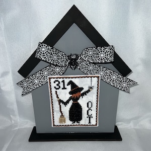 May include: A gray wooden house-shaped frame with a black and white spiderweb ribbon bow. The frame holds a cross-stitch design of a witch with a broom and a spider hanging from a web. The design also includes the text "31 OCT".