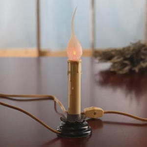 May include: A cream-colored electric candle lamp with a flame-shaped bulb. The candle has a dripping wax effect and sits in a black base. A brown cord and plug are attached. The lamp is on a dark brown surface.