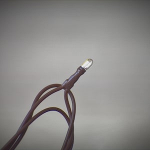May include: Close-up of a single LED light bulb with brown wires. The bulb is clear and emits a bright light. The wires are looped and appear to be part of a string light set. The background is a solid, neutral gray.