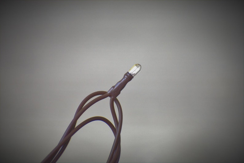May include: Close-up of a single LED light bulb with brown wires. The bulb is clear and emits a bright light. The wires are looped and appear to be part of a string light set. The background is a solid, neutral gray.