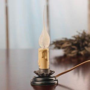 May include: A vintage-style electric candle lamp with a cream-colored, wax-like body and a clear, flame-shaped bulb. The lamp sits on a black base with a decorative metal holder and has a brown electrical cord.
