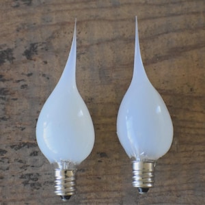 May include: Two white, teardrop-shaped light bulbs with a pointed tip. The bulbs have a frosted appearance and are set against a wooden background. The base of each bulb is silver-colored.