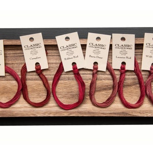 May include: Six spools of embroidery floss in various shades of red, labeled with the names "Holly Berry", "Campfire", "Ribbon Red", "Barn Door", "Licorice Red", and "Wild Berries".